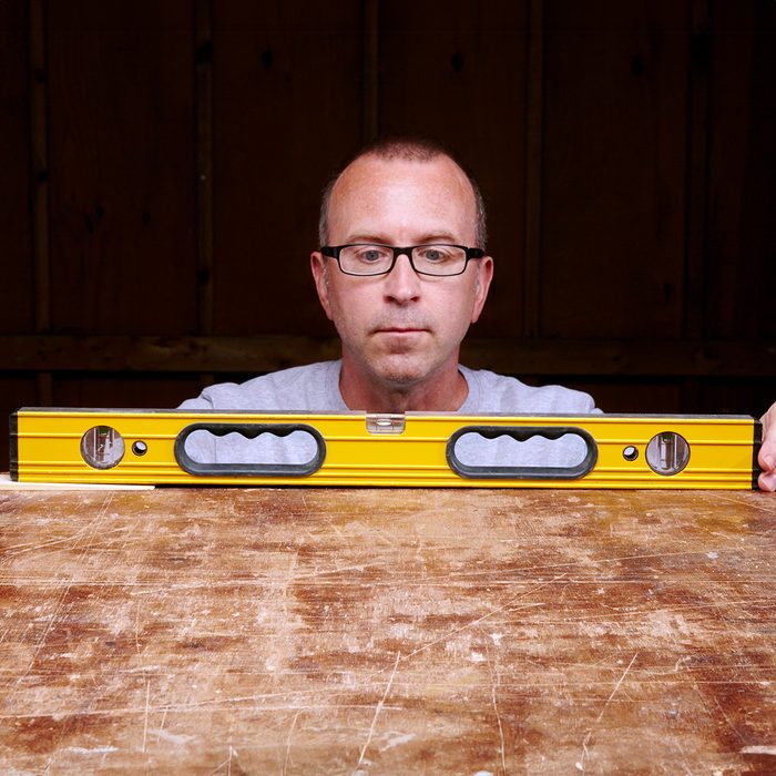 A person wearing glasses is looking at a yellow spirit level placed on a wooden table, ensuring it is level. The background is dark and wooden.
