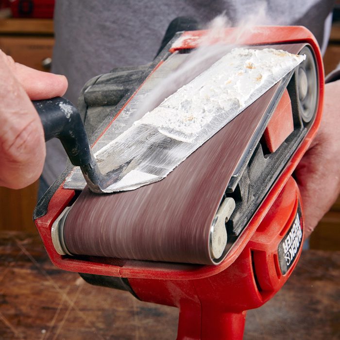 A person using a red belt sander to smooth a piece of metal, with dust being generated. The sander is held firmly, and the metal shows signs of sanding. Tools are on a wooden surface in the background.