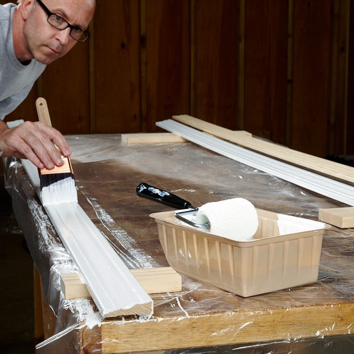 A person wearing glasses is painting a piece of wood molding white on a table covered with plastic. A paint brush, roller, and tray are nearby. The background has wood-paneled walls.