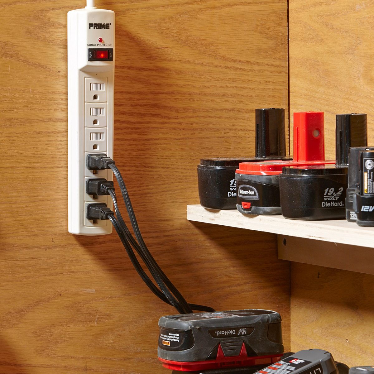 A white power strip mounted on a wooden wall has multiple black cords plugged in. Nearby, several black and red battery packs and power tool accessories are neatly arranged on a shelf.