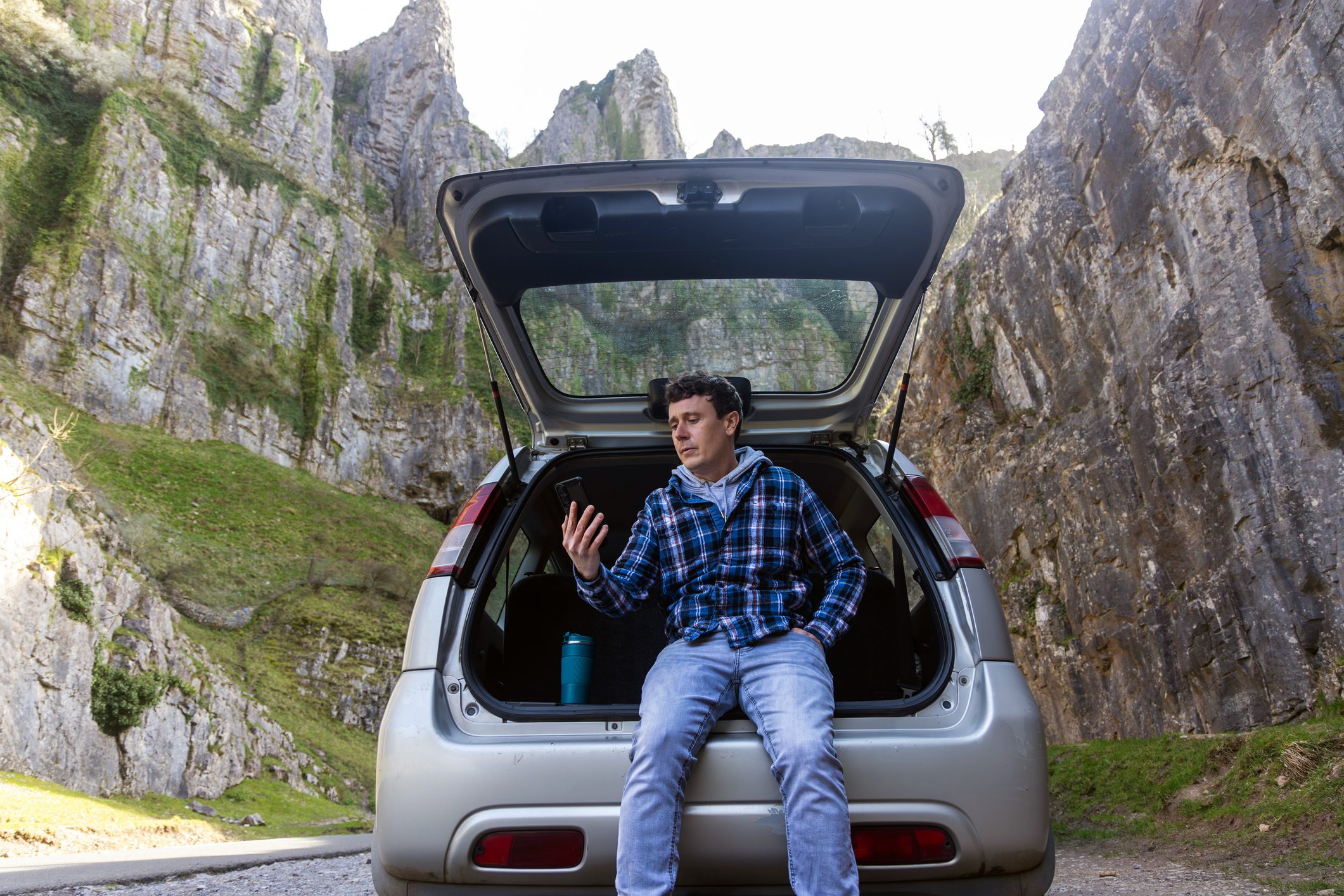 Man sitting on edge of car trunk watching a video on his phone, with mountains in the background