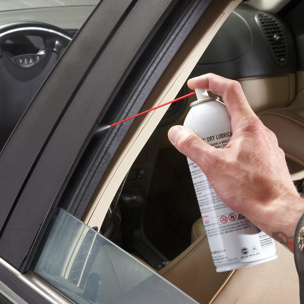 A person uses a can of spray lubricant with a narrow straw attachment to apply lubricant to the window channel of a car door. The car window is slightly open, and the interior of the car is visible.