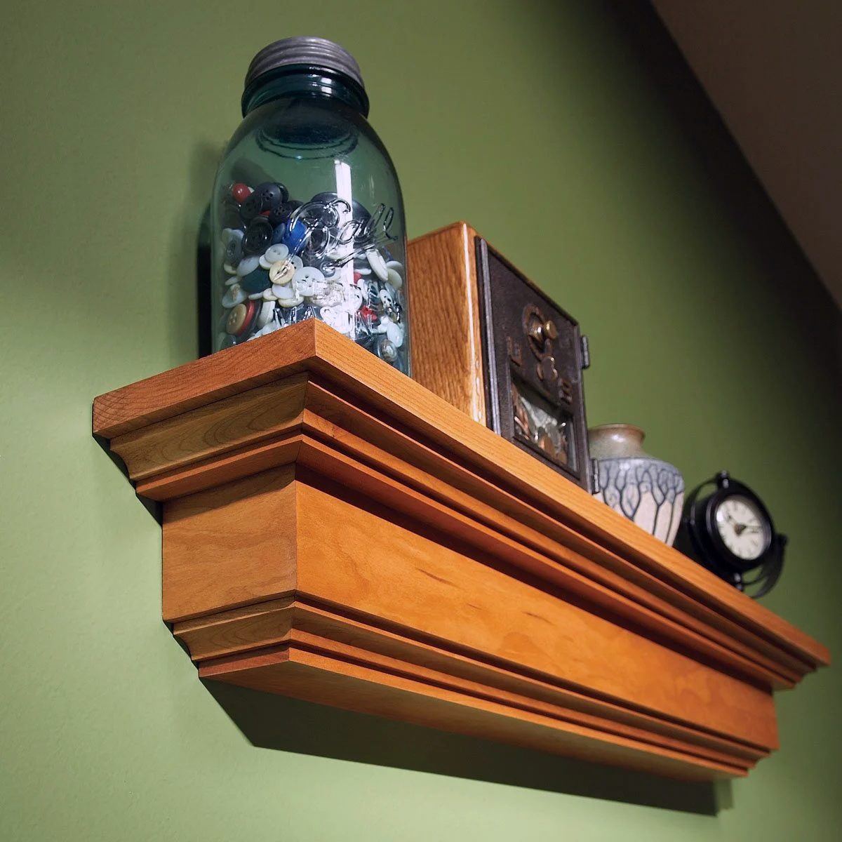 A wooden shelf mounted on a green wall displays a blue jar filled with small items, a wooden box, a ceramic vase with a blue pattern, and a black clock.