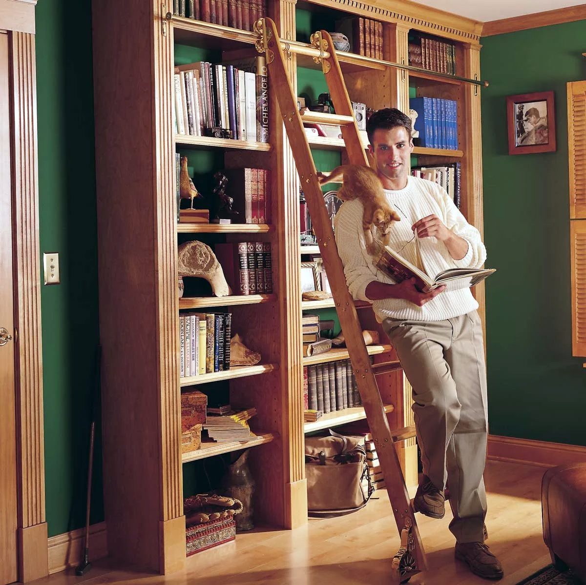 A person stands on a ladder in front of a large wooden bookshelf, reading a book. The shelf is filled with various books and decorations. The room has green walls and wooden flooring.