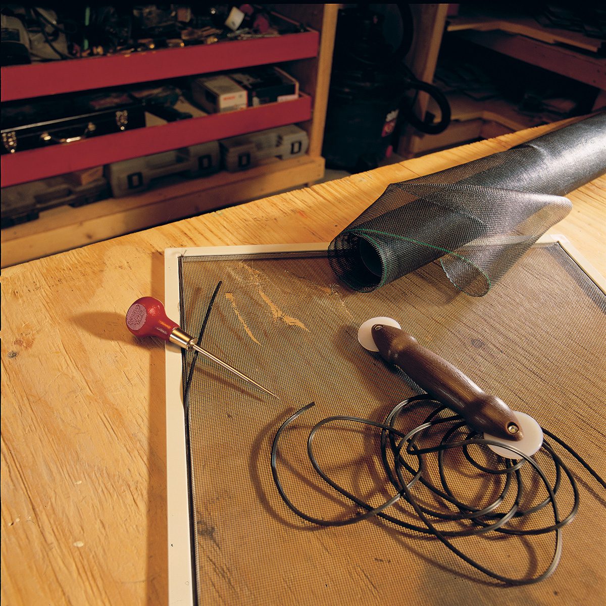 A workbench with a wooden surface holds a small red-handled tool, thin black wires, a sheet of material, and other small parts. Shelves with various tools and items are visible in the background.