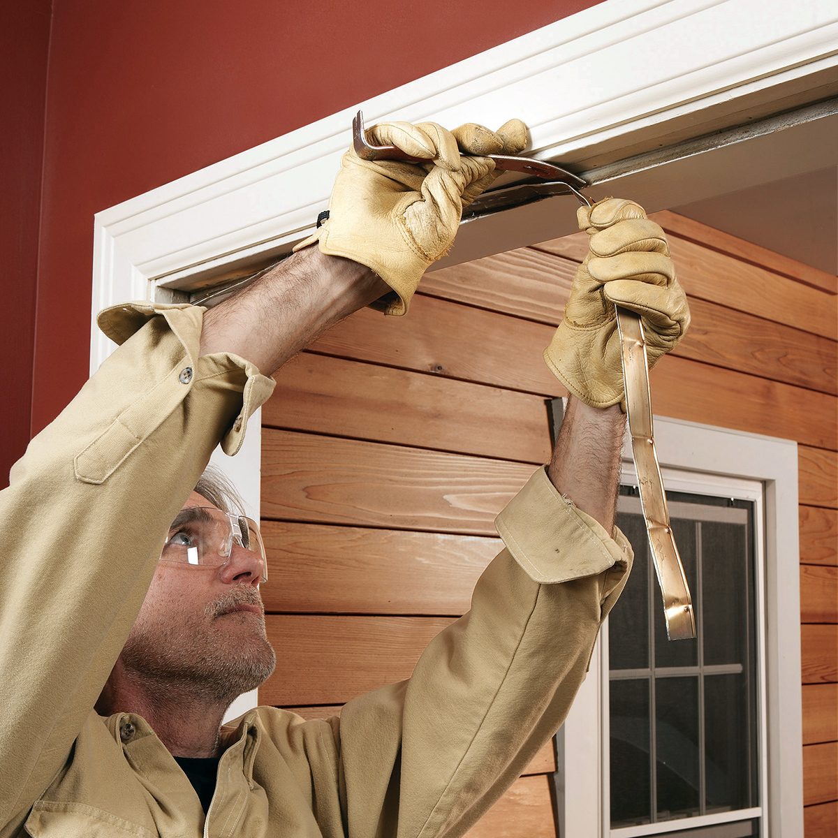 A person wearing safety goggles and gloves uses a pry bar to remove trim from a wooden door frame. The wall is wooden with a window beside the door. The person is focused on the task.