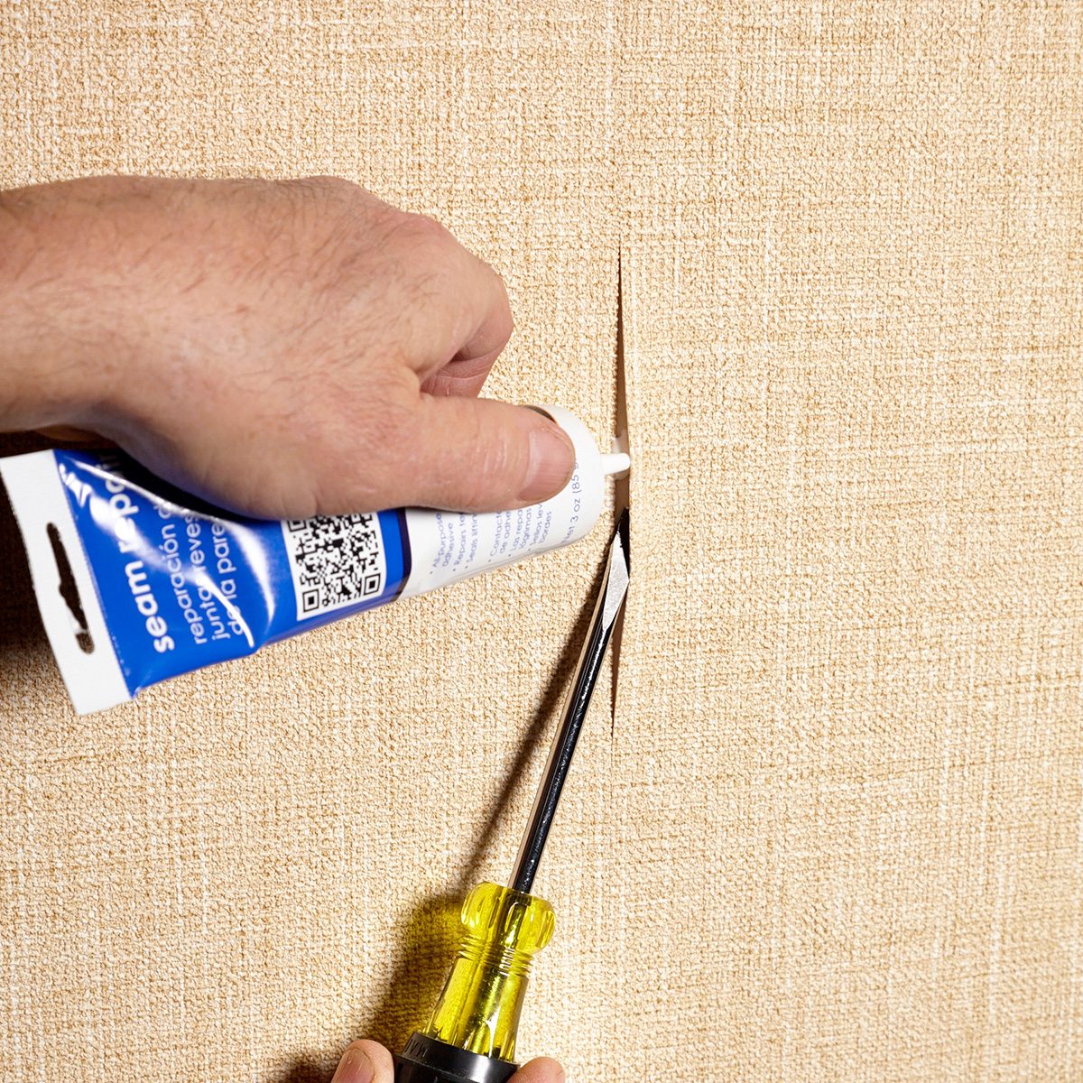 A person is repairing a vertical tear in beige wallpaper using a tube of seam adhesive. They hold the tube with one hand while using a screwdriver to open the tear slightly for easier application.