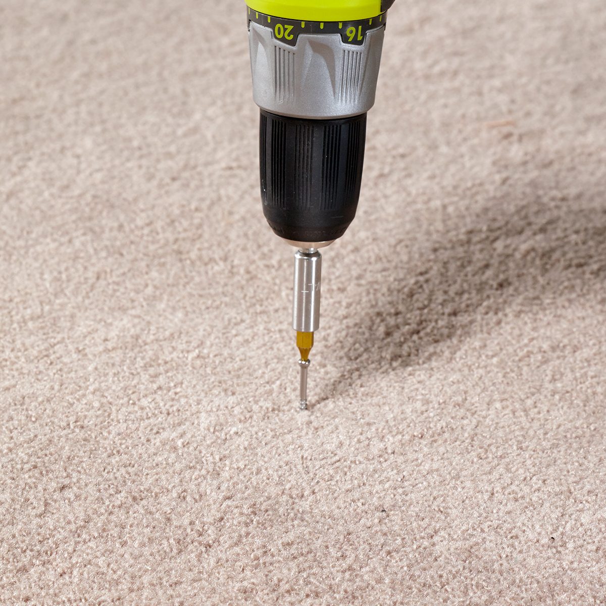 A power drill with a screwdriver bit is positioned downward onto a beige carpeted surface. The drill has a black and green grip, and the bit is partially pressed into the carpet.