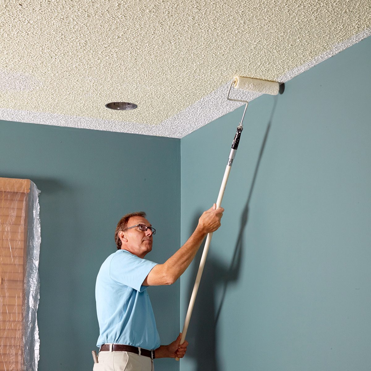 A person in a blue shirt is using a long-handled roller to paint a ceiling beige in a room with teal walls. A covered object is partially visible in the corner.