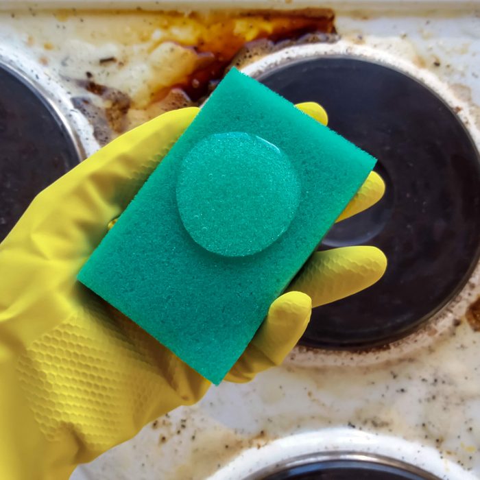 hand in household glove holding sponge with cleaning liquid over dirty stove