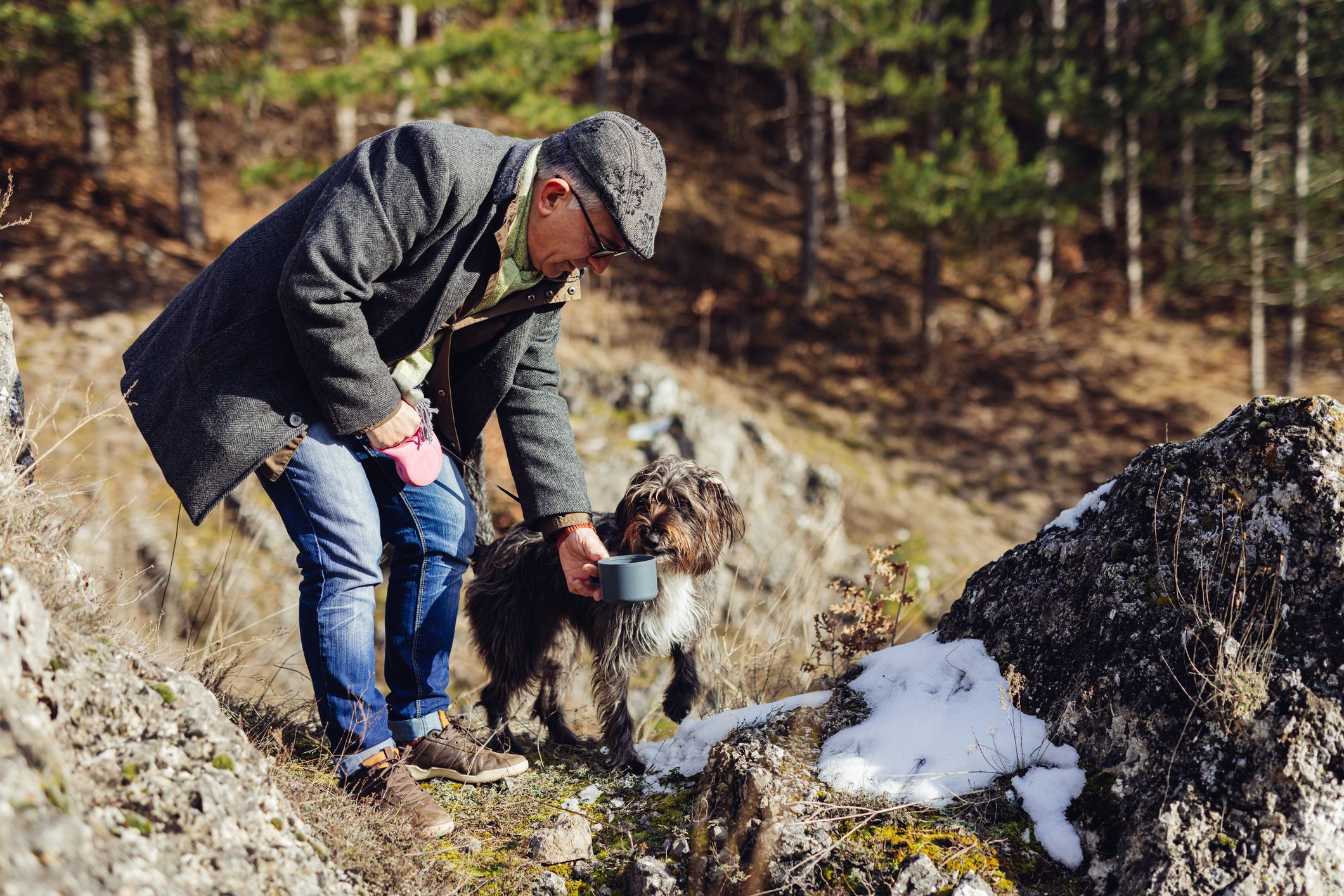 Senior man gives water to his dog while out for a walk in the woods in winter