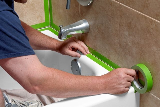 A person applies painter’s tape around the edges of a bathtub and fixtures, preparing to caulk or paint, in a bathroom with tan tiled walls.