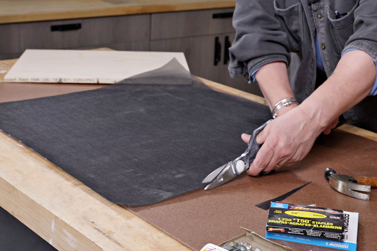 a person is cutting a black textured material with scissors; the material is laid flat on a wooden table next to a piece of light colored wood; various tools and supplies are visible, including a hammer and a box of staples;