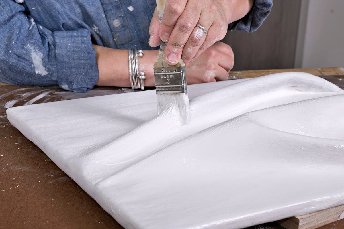 A person is seen applying white paint to a textured surface using a brush; the individual is wearing a denim shirt that appears to have specks of white paint on it; with their hand holding the brush firmly;