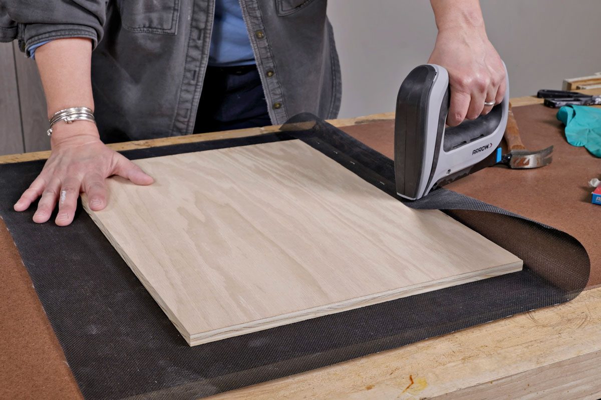 a person's hand holding an electric stapler, pressed against a piece of wood placed on a work surface; The wood appears to be a light colored panel, plywood, and is resting on a dark mesh fabric; Other tools and items are visible on the workbench, including a pair of green gloves, a hammer, scissors, and a box of staples;