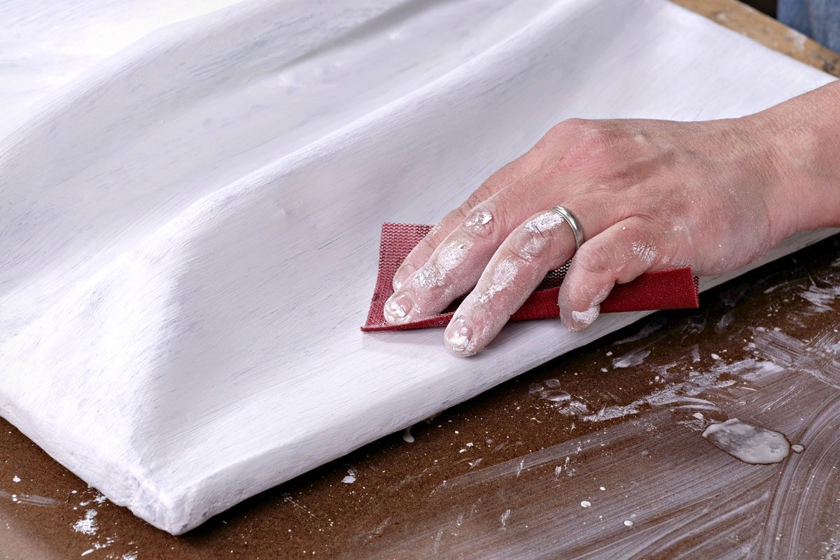 a hand engaged in sanding a textured white surface with a piece of sandpaper, the hand appears to be covered in a layer of white dust, the workspace underneath seems to be made of a flat, brown material