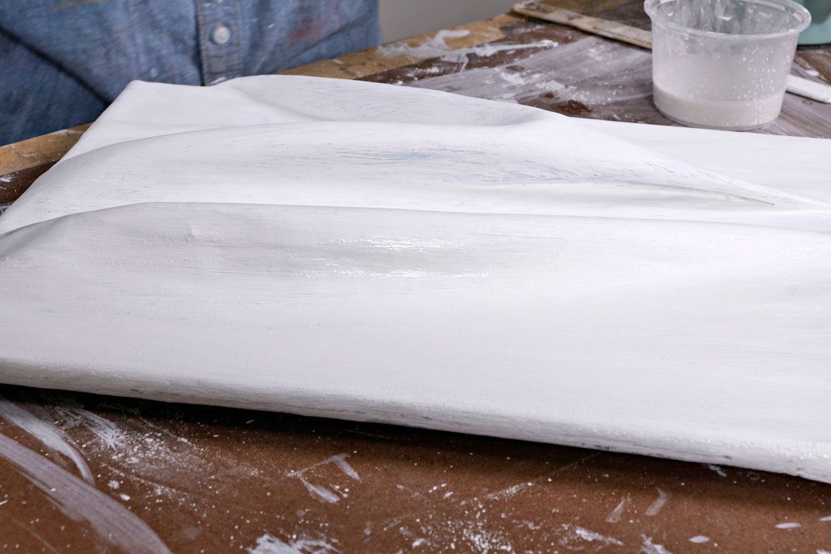 a large, textured white surface that appears to be an art canvas or a piece of wood coated with a white primer; in the background, a work surface with scattered traces of white paint, a small container filled with paint or a similar substance