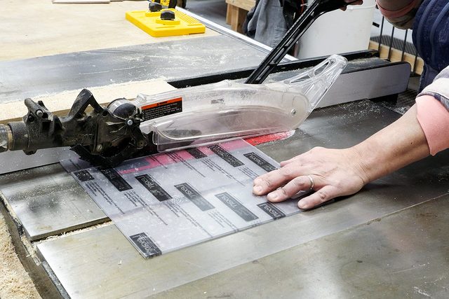 A person uses a table saw with a clear blade guard to cut a laminated sheet, keeping hands safely away from the blade in a woodworking shop.