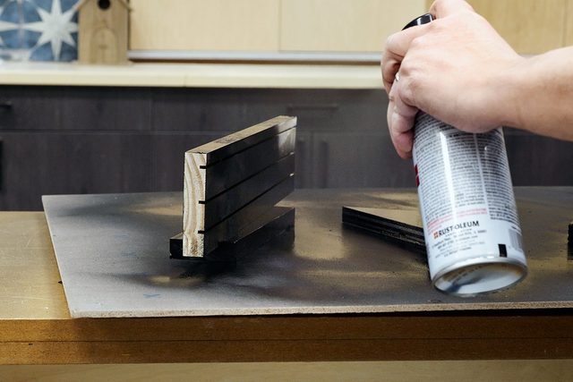 A person sprays black paint from a can onto a small wooden object placed on a table, with protective paper underneath to catch the excess paint.