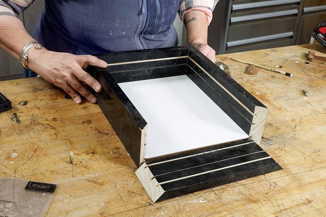 A person is assembling a rectangular picture frame with black sides on a wooden workbench. The frame corners are mitered, and tools are scattered in the background.