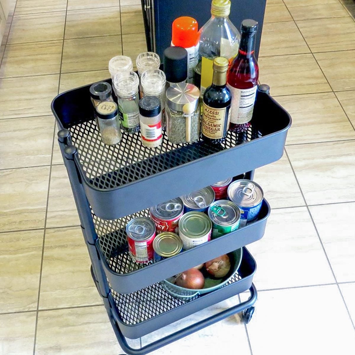 A black rolling utility cart with three shelves holding spices, condiments, canned drinks, and a bowl with onions, standing on a tiled floor.