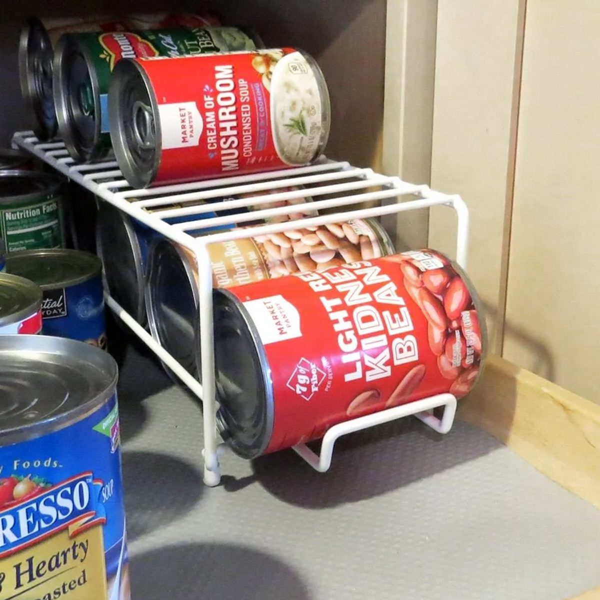 Cans of food, including light red kidney beans and cream of mushroom soup, are organized on a white metal rack inside a kitchen cabinet. Some other cans are visible to the side and underneath the rack.