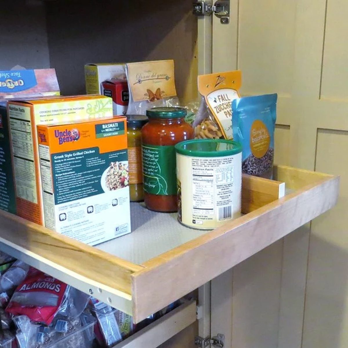 A kitchen pantry with a pull-out wooden shelf holding various food items, including cereal boxes, a jar of pasta sauce, a can, and bags of snacks or dry goods. The pantry door is open.