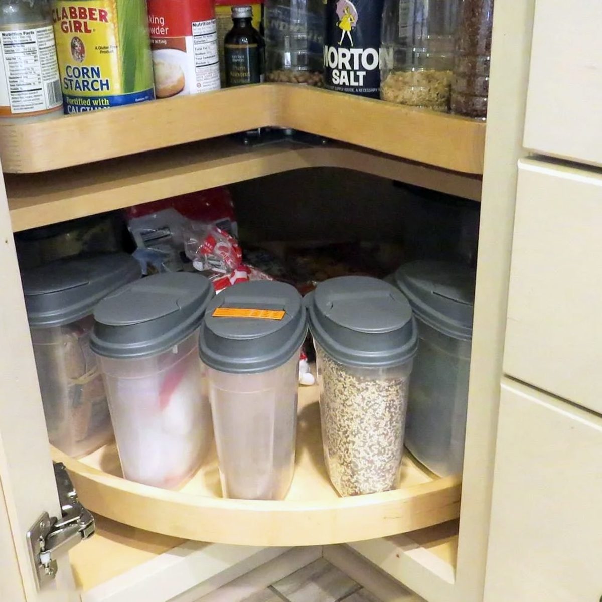 A kitchen corner cabinet with two rotating shelves, holding various pantry items. The top shelf has canned and bottled goods, while the bottom shelf has plastic containers filled with dry foods.
