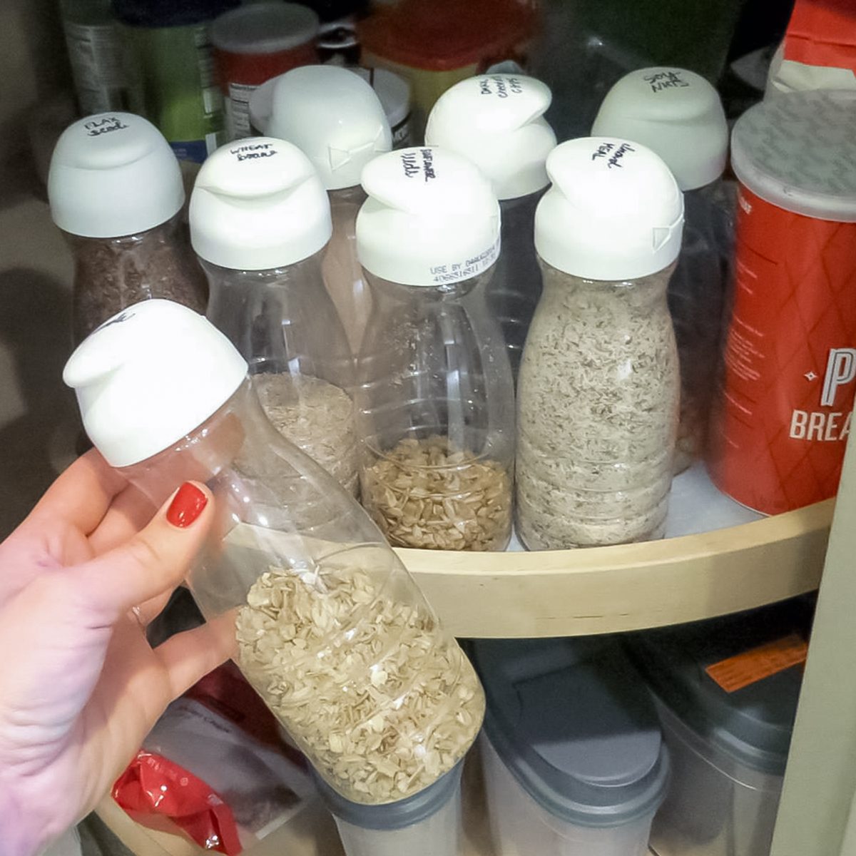 A hand holds a plastic bottle filled with oats over a rotating pantry shelf containing several similar bottles with white lids, each filled with different dry foods.