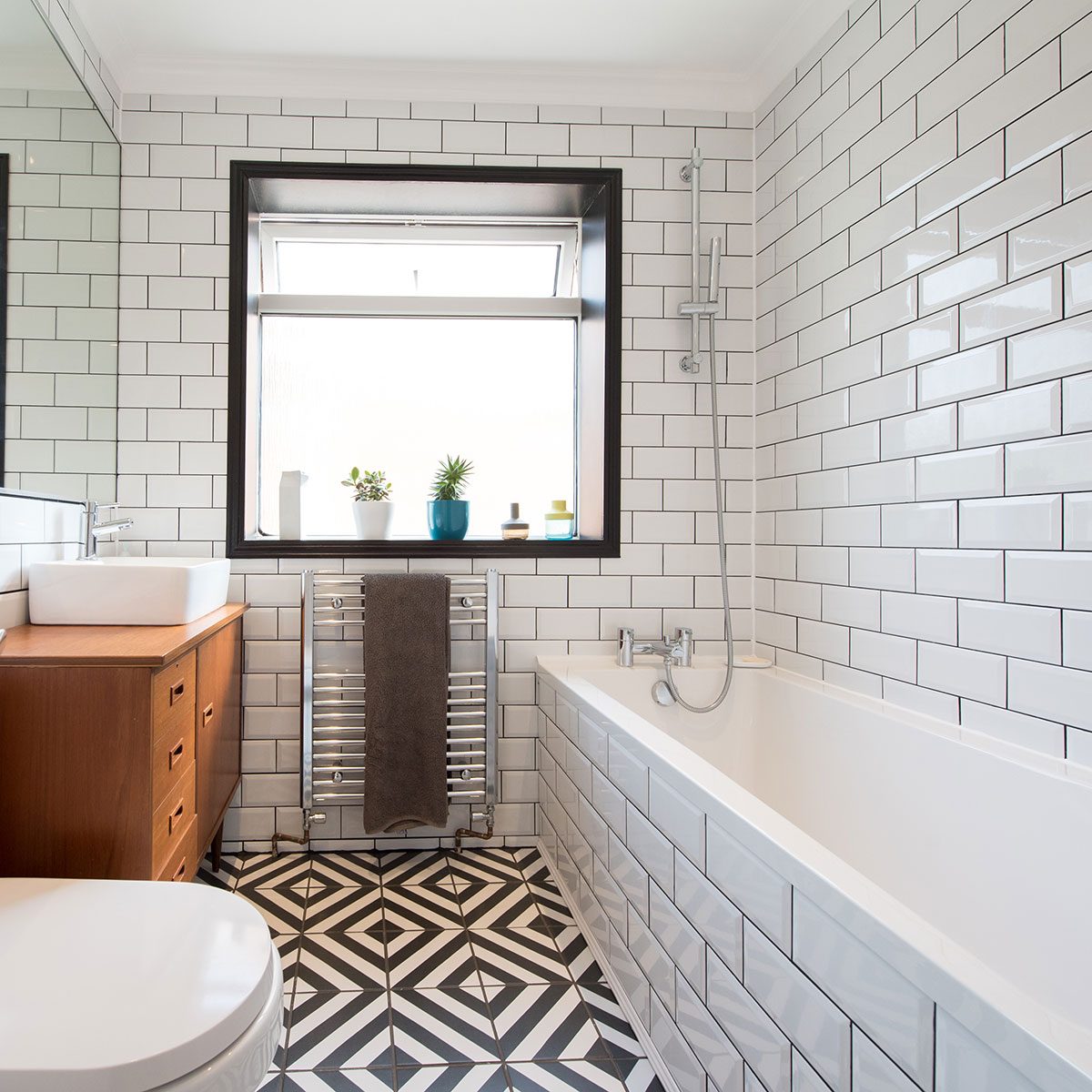 A general view of a modern bathroom with a square sink sitting on a wooden dresser with chrome tap, potted plants on the window sill, towel rail radiator, bath, black and white tiled floor and large mirror.