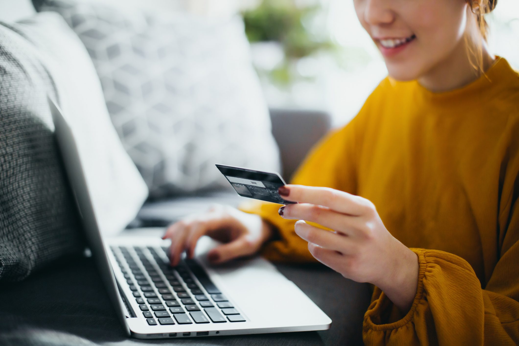 Close up of beautiful smiling young woman sitting on the floor by the sofa, managing financial bill payment with laptop and credit card on hand at cozy home. Technology makes life so much easier