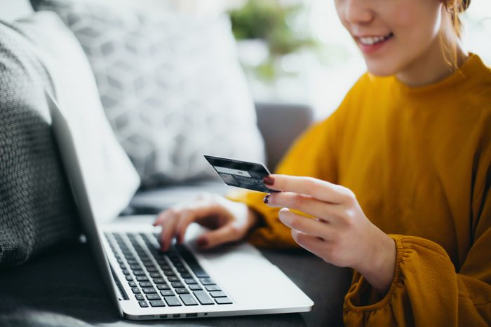 Close up of beautiful smiling young woman sitting on the floor by the sofa, managing financial bill payment with laptop and credit card on hand at cozy home. Technology makes life so much easier