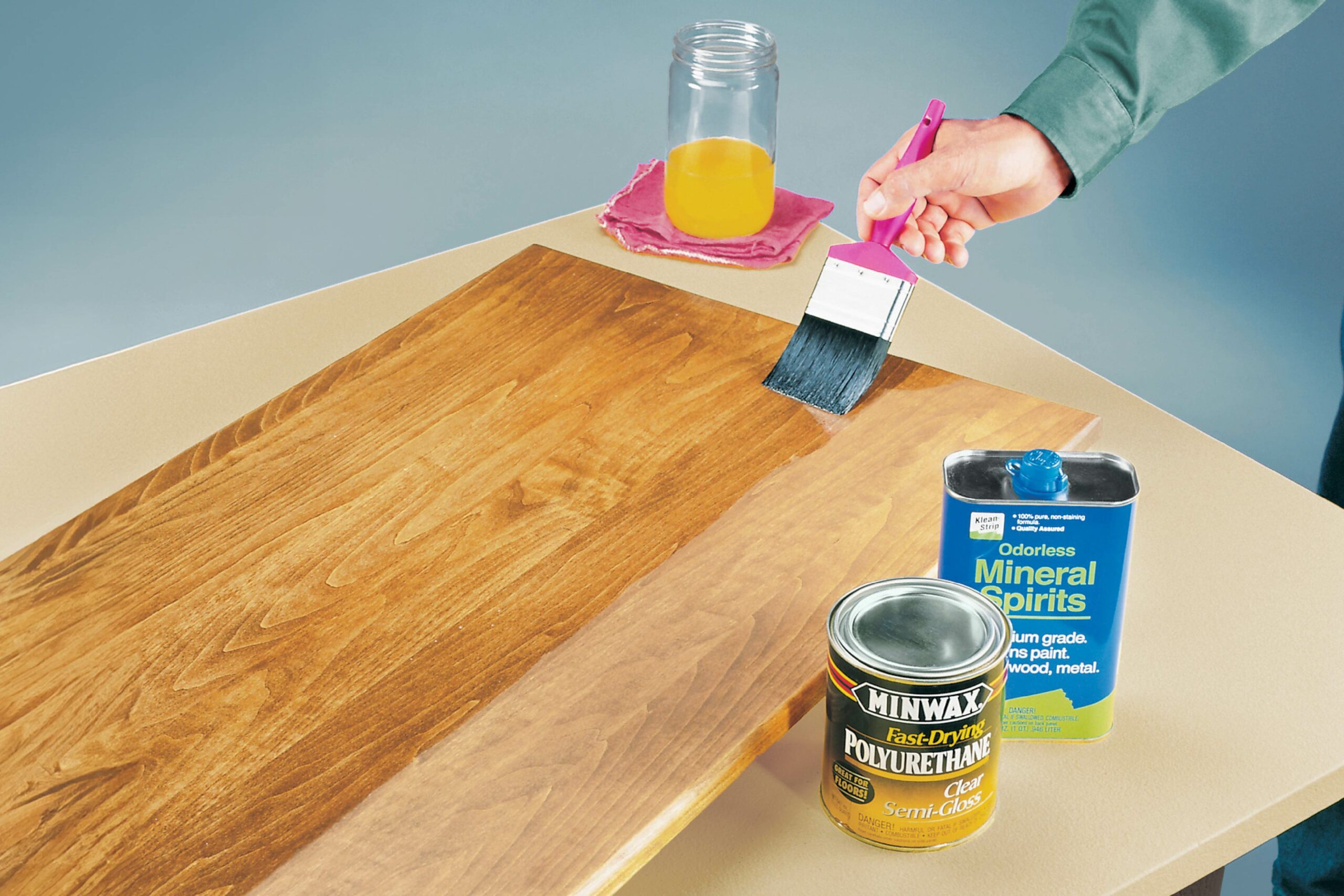 A person brushes polyurethane onto a wooden surface on a brightly colored table, with mineral spirits and a jar of liquid nearby.