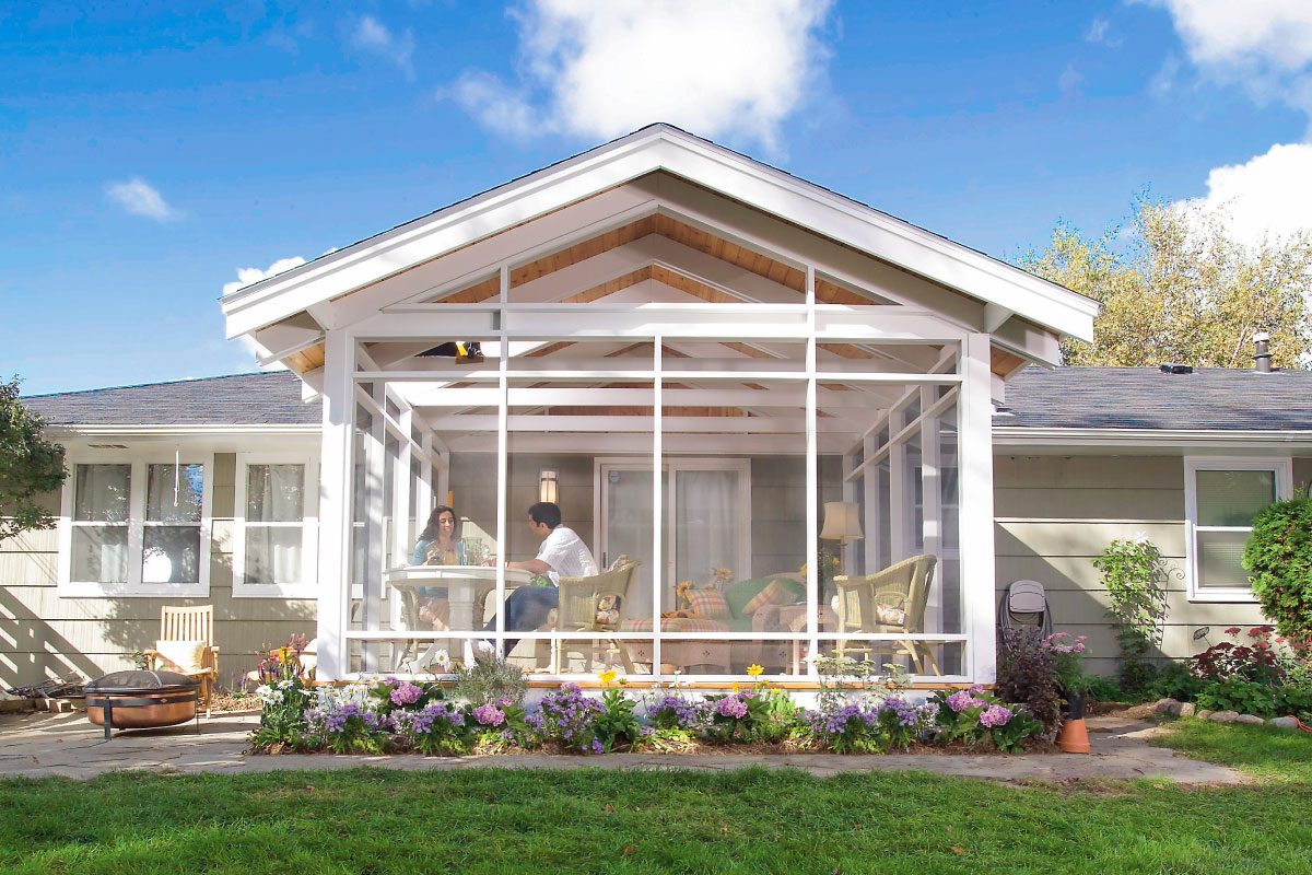 A sunroom features two people sitting at a table, engaged in conversation, surrounded by colorful flowers and green grass on a clear day.
