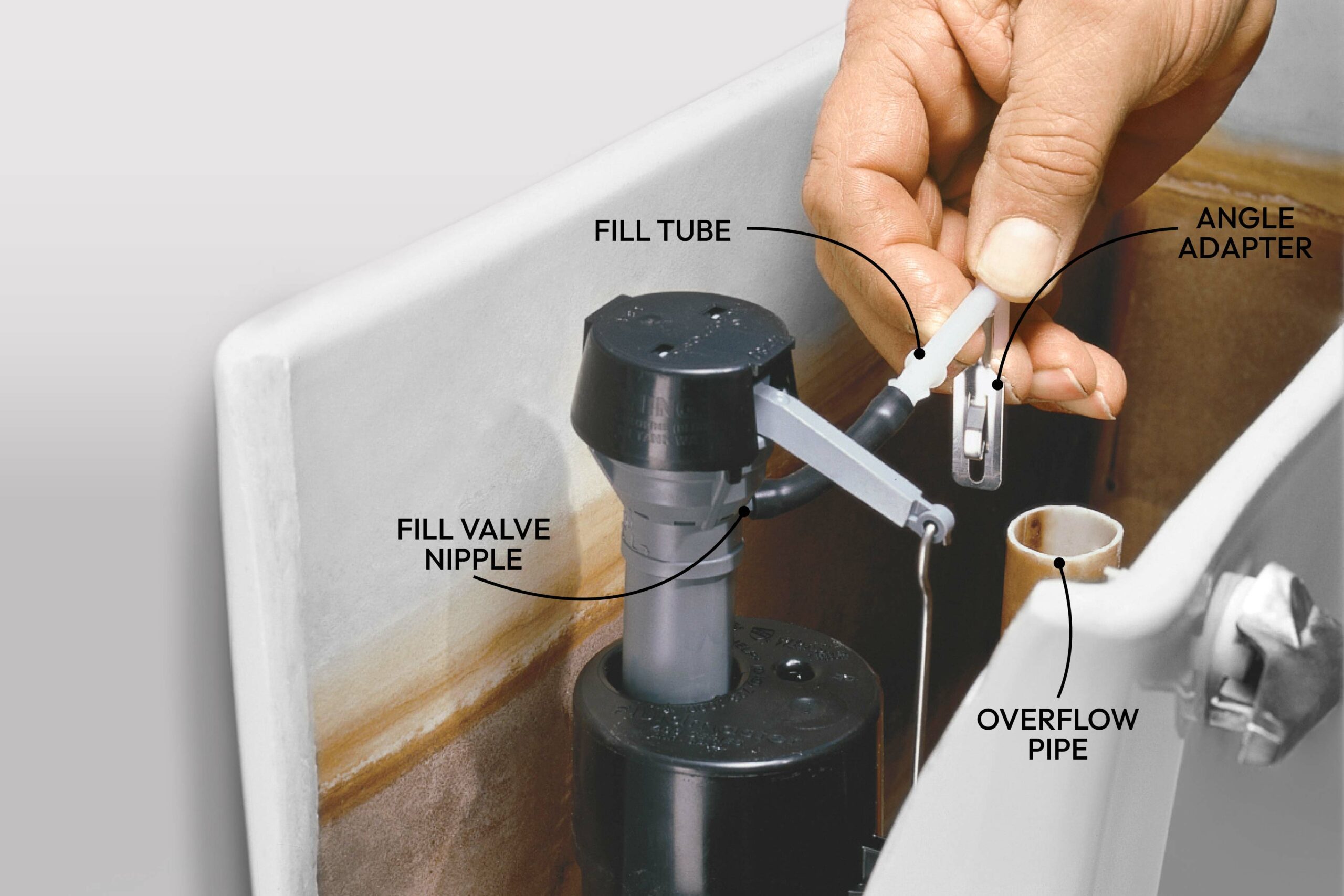 A hand connects a fill tube to a fill valve nipple inside a toilet tank, surrounded by various plumbing components including an overflow pipe and angle adapter.