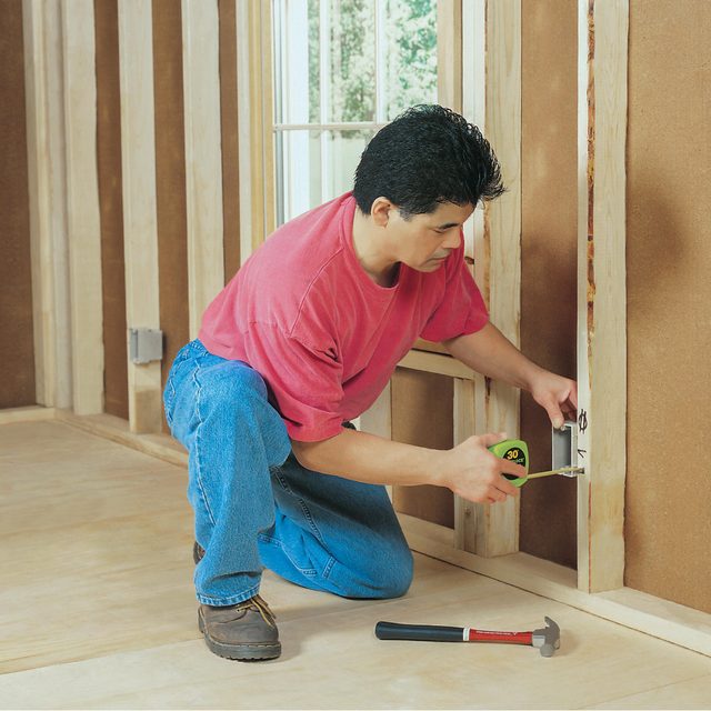 A man kneels while measuring a wall with a tape measure, surrounded by wooden framing in a construction area. A hammer lies beside him.