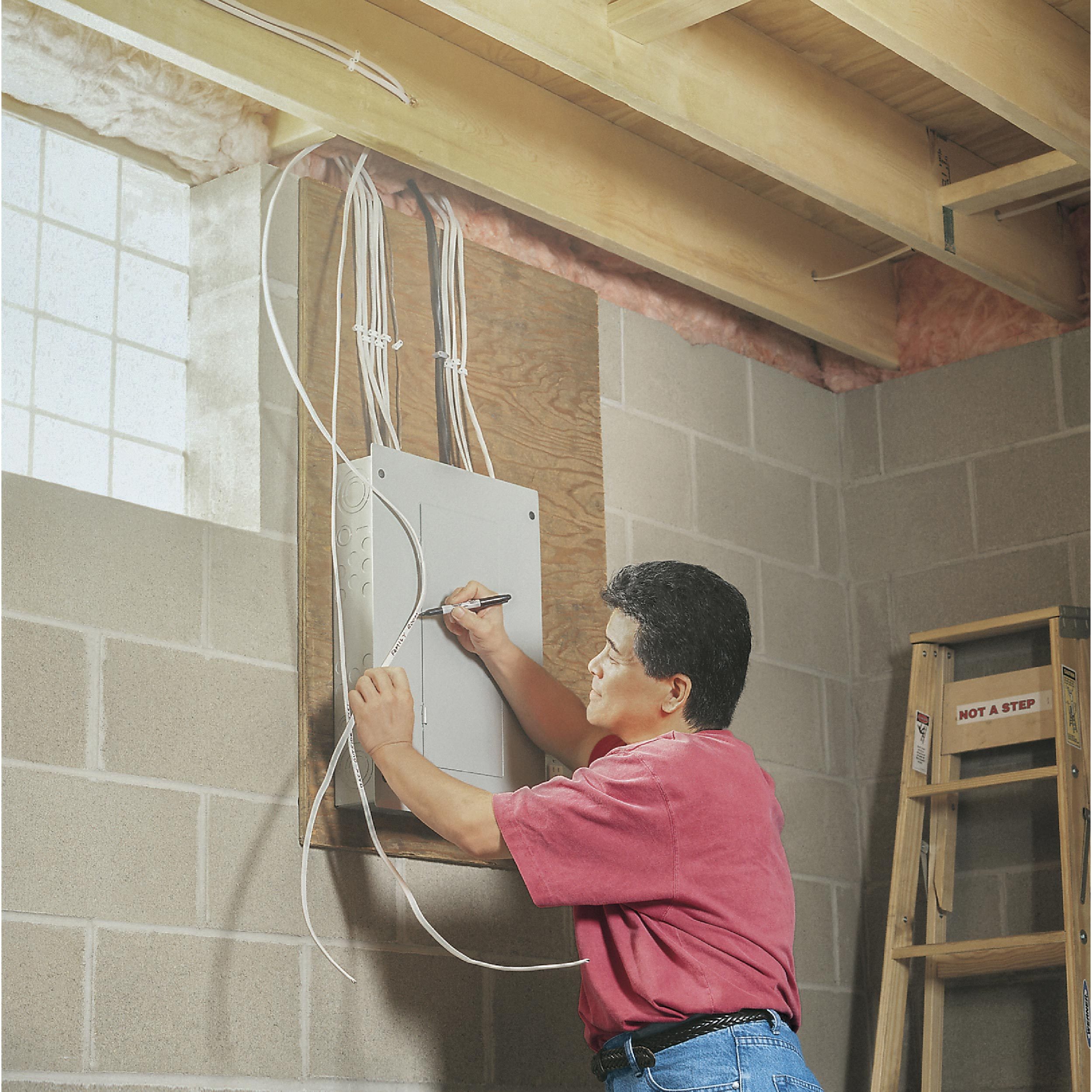 A man installs and labels wires on an electrical panel in a basement, surrounded by gray cinderblock walls and wooden framing.