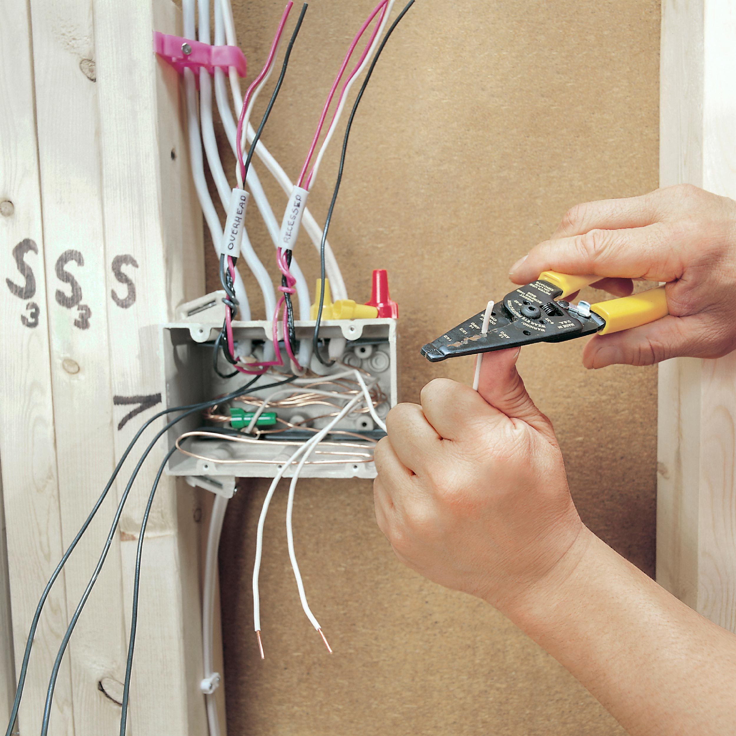 A person's hand uses a wire stripper on a copper wire, while various insulated wires connect to an electrical box mounted on a wall.