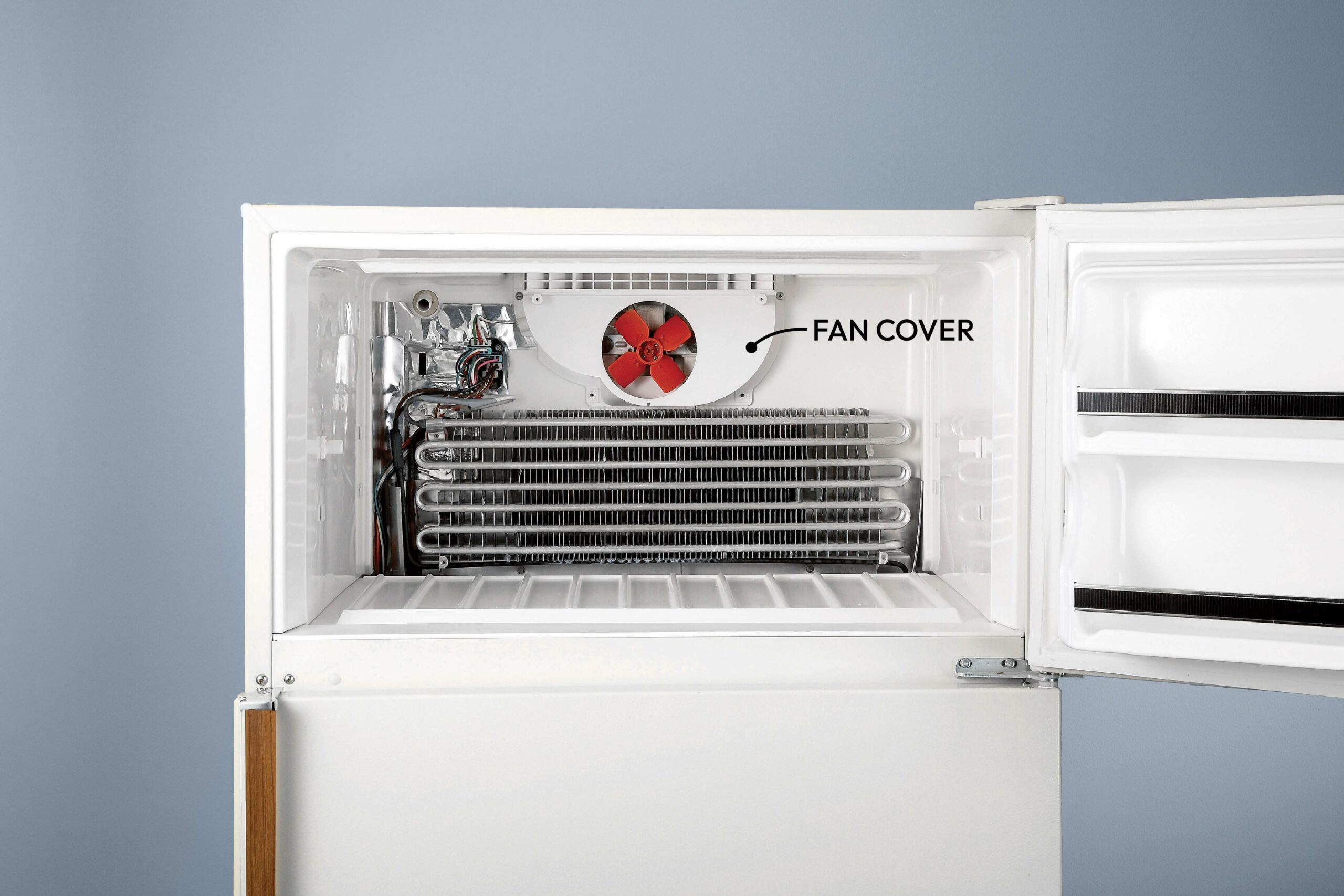 An open refrigerator interior features a red fan behind a cover and metal coils for cooling, set against a grey wall.