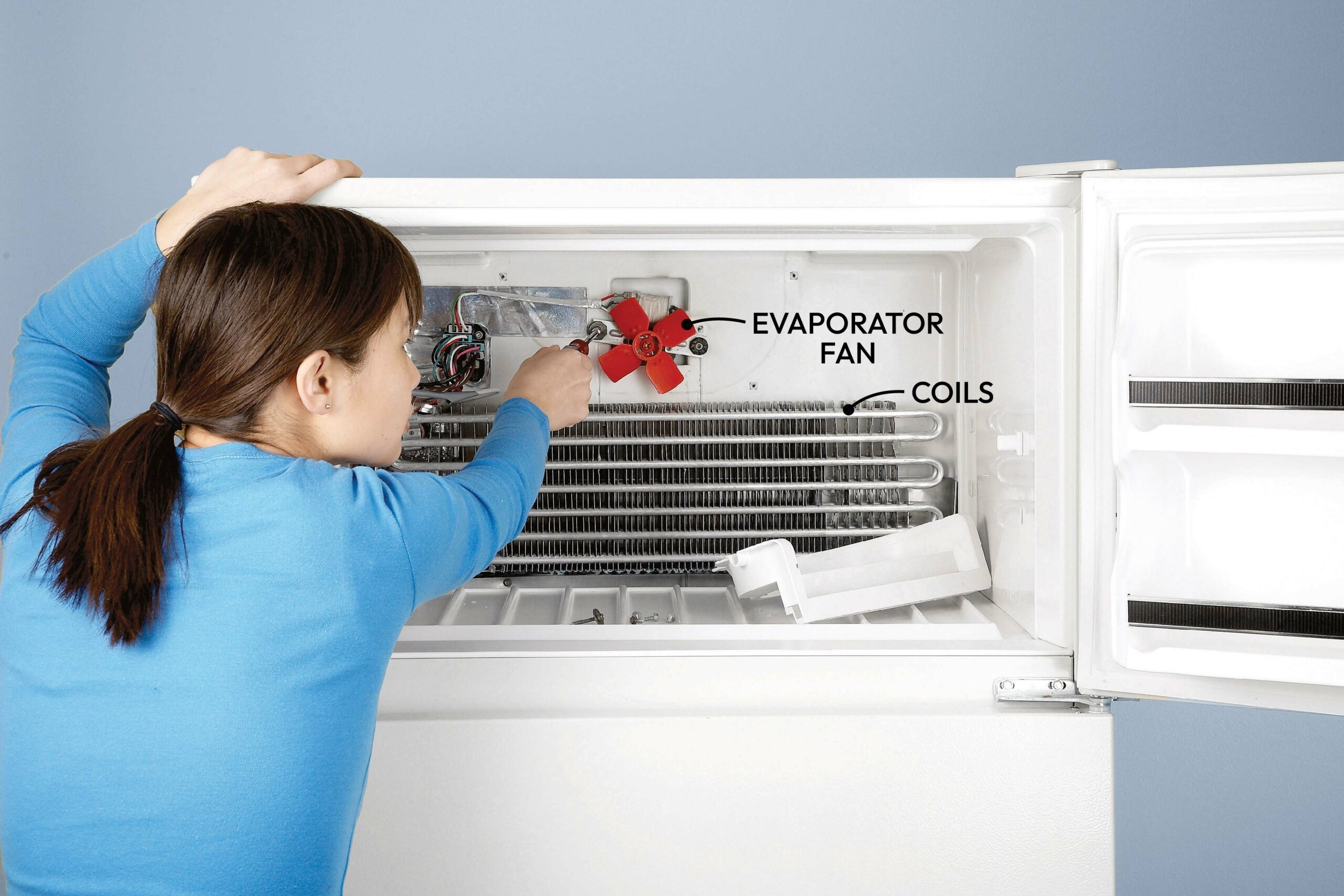 A person is repairing an evaporator fan in a freezer, accessing internal components like coils, with tools and parts visible in the interior space.