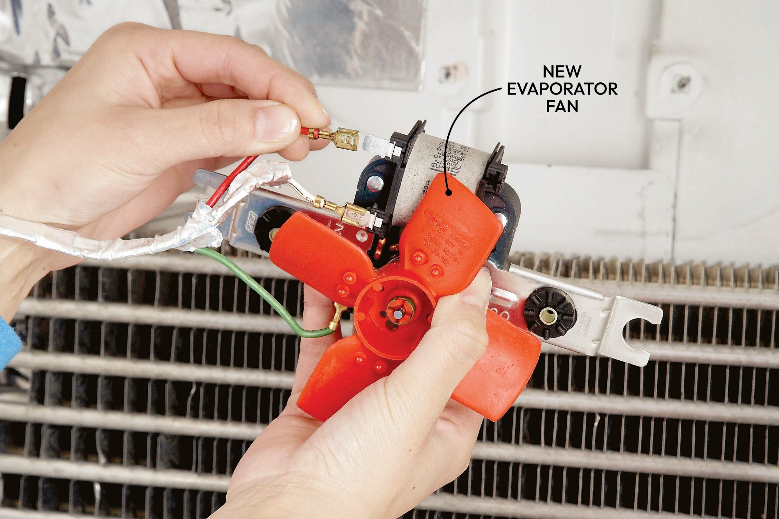 A person holds a red evaporator fan, connecting wires, while an air conditioning unit's cooling fins are visible in the background.