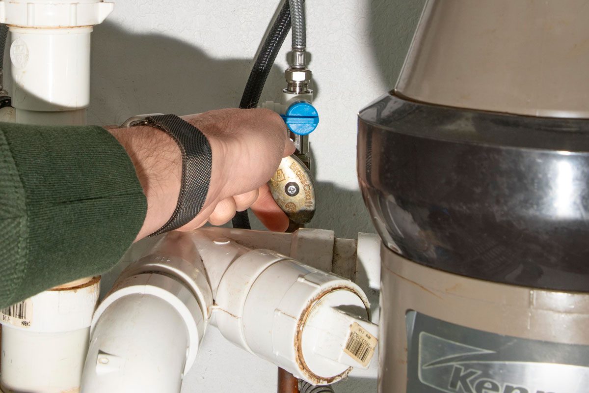 a person's hand turning off a water valve under a sink, the valve has a blue handle and a brass body, there are white PVC pipes and a black flexible hose connected to the valve, a garbage disposal unit labeled Kenmore is visible on the right side of the frame, the wall behind the plumbing is off white