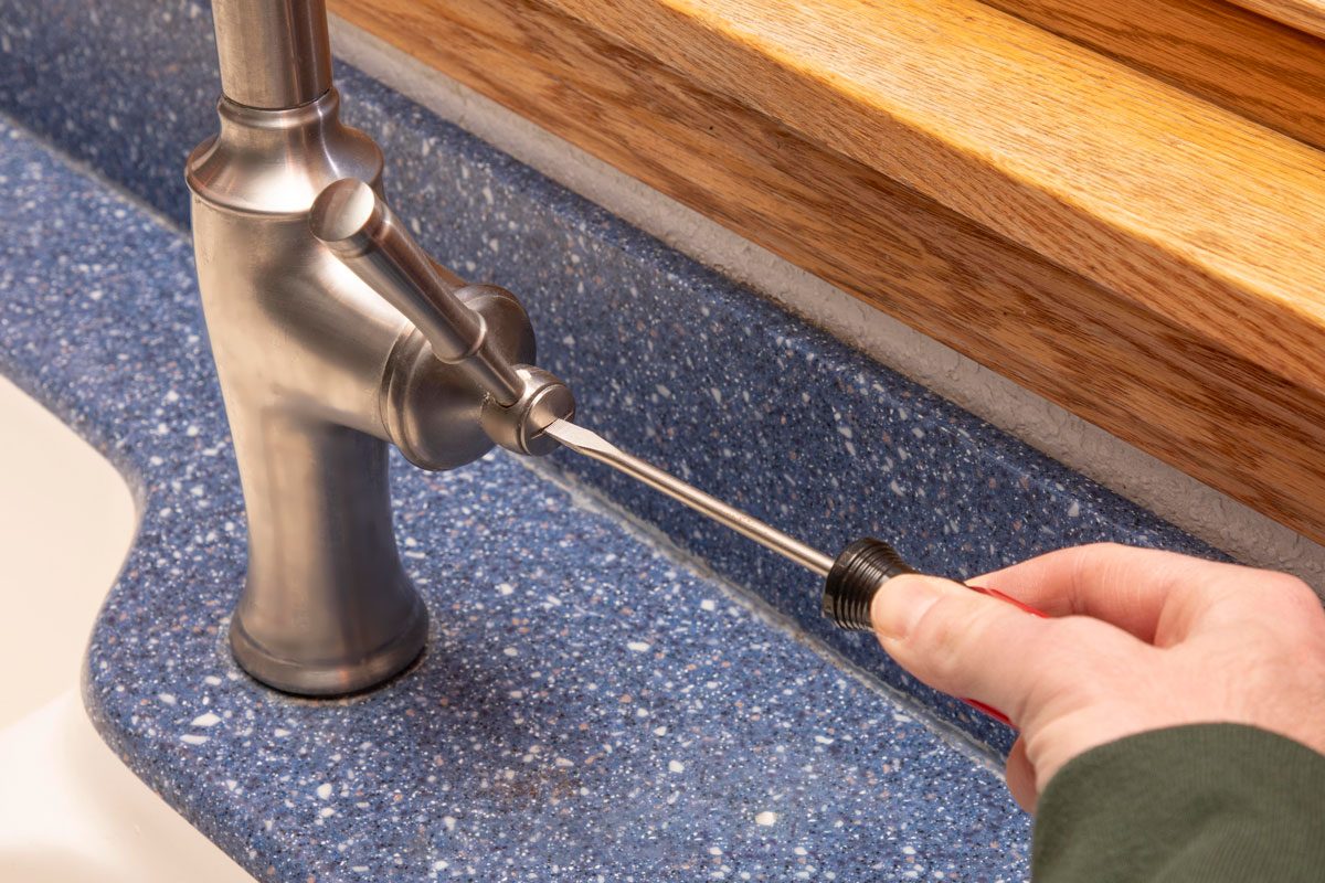 a hand holding a screwdriver and working on the side of a brushed metal kitchen faucet; the faucet is mounted on a blue speckled countertop with a white sink basin visible to the left; a wooden window sill is visible in the background