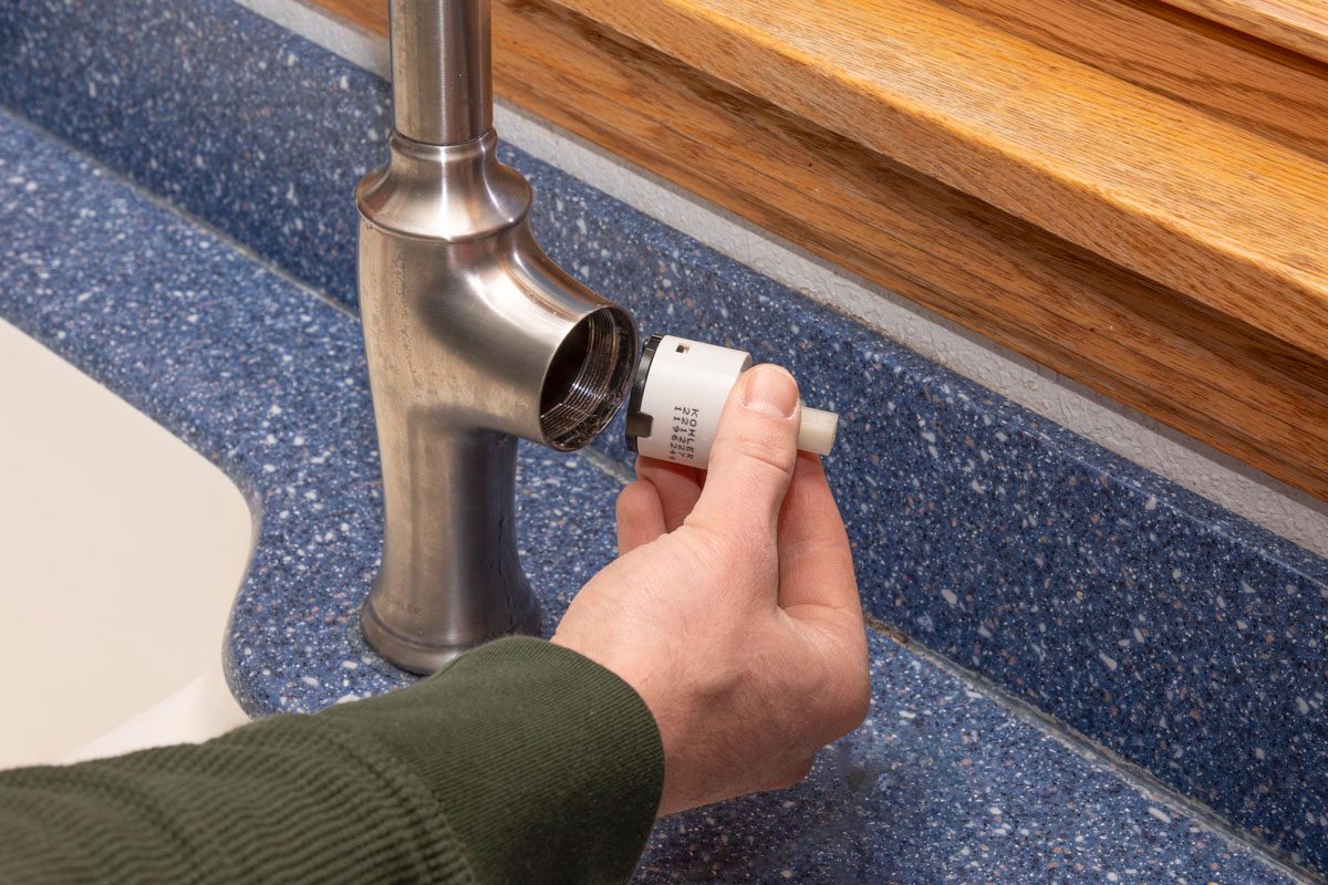 a hand inserting a white cylindrical cartridge into the side of a brushed metal kitchen faucet; the faucet is mounted on a blue speckled countertop, and a white sink is partially visible on the left; a wooden window sill is in the background