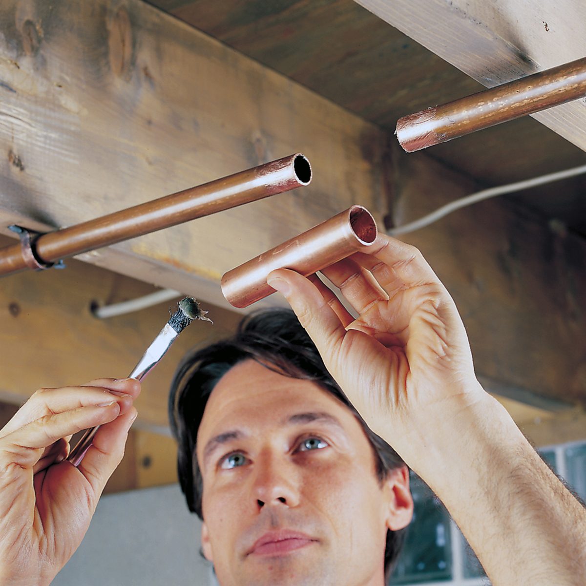 A man applies flux to the end of a copper pipe with a brush, preparing to join it to another pipe under a wooden ceiling.