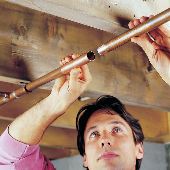 A person is fitting two copper pipes together on the ceiling, appearing to work on plumbing installation or repair in an unfinished space with exposed wooden beams.