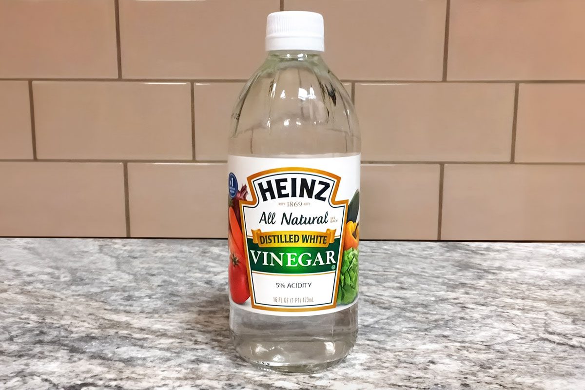 A clear plastic bottle of Heinz All Natural Distilled White Vinegar sits on a gray marbled countertop with a beige tiled wall in the background;