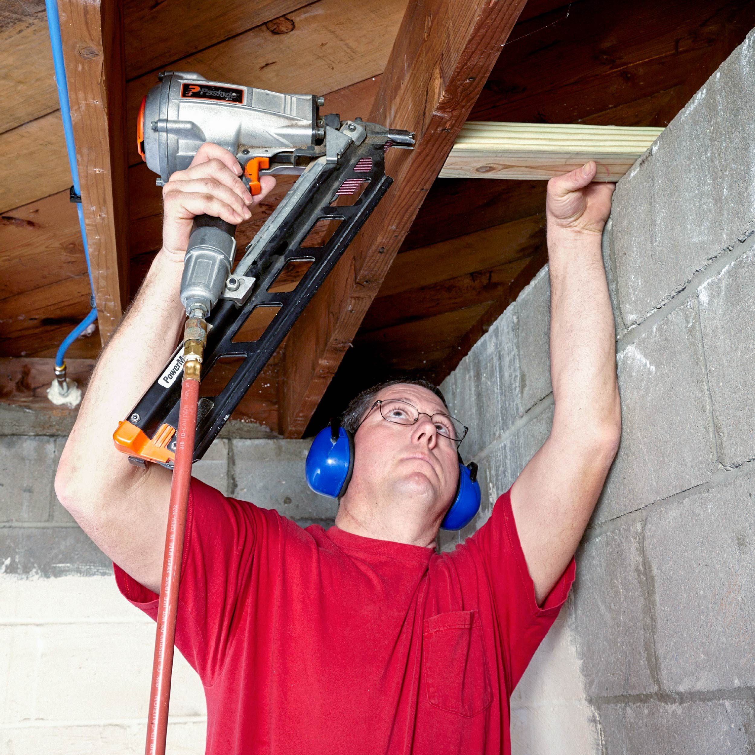 A man uses a nail gun to fasten a wooden beam in a basement, surrounded by concrete walls and wooden beams above.