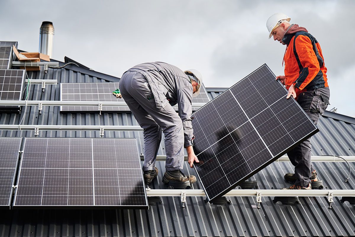 Technicians carrying photovoltaic solar module while installing solar panel system on roof of house