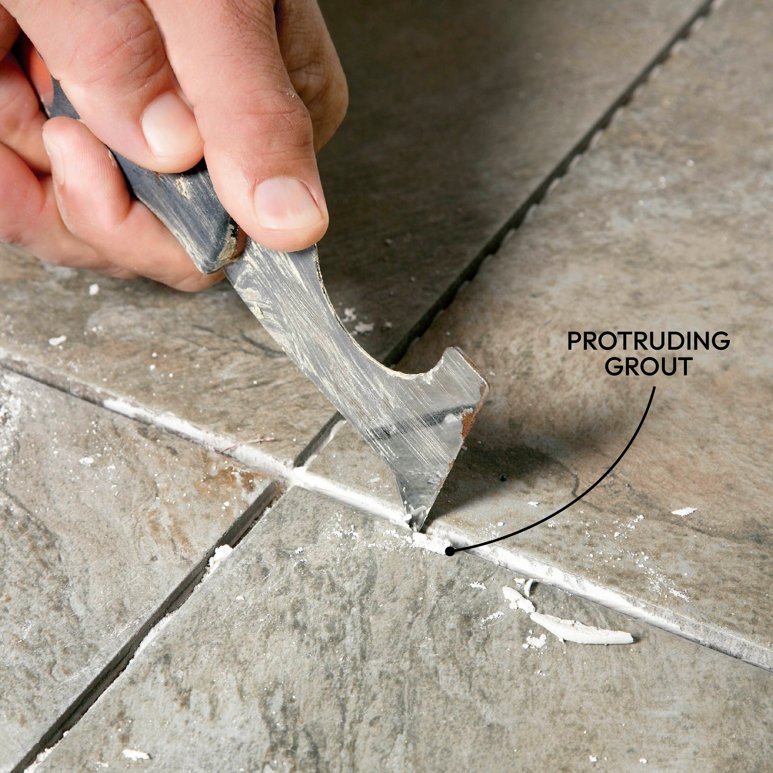 A hand holds a tool, scraping excess grout from between ceramic tiles on a floor, with dust and debris visible in the surrounding area.